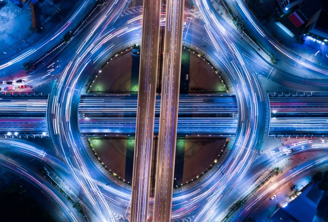 Aerial long-exposure photograph of a circular highway interchange at night, with streams of blue, white, and gold light trails illustrating the complexity and interdependence of critical infrastructure systems.