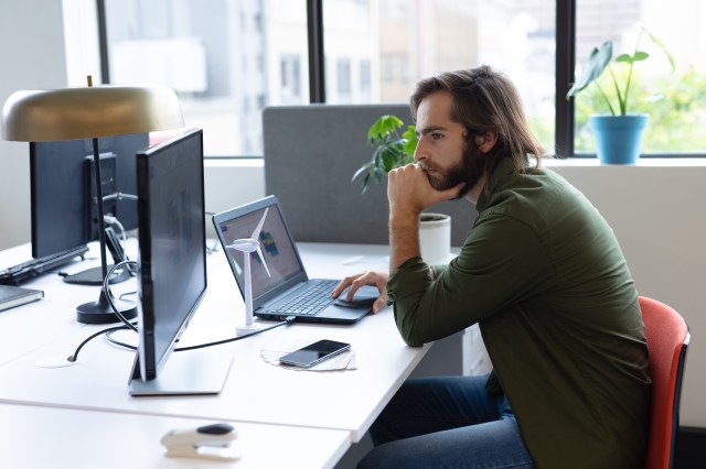 A professional reviews data on a monitor at a modern office desk, leaning forward in a posture of focused concentration.