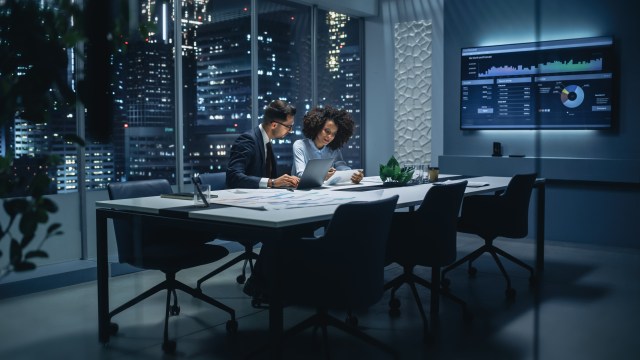 Two business professionals reviewing cybersecurity risk reports in a boardroom with a data dashboard on screen