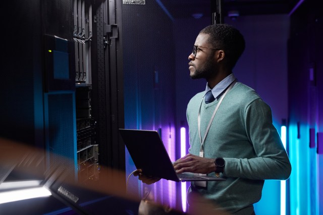 An IT engineer wearing a lanyard reviews data on a laptop while standing in front of server hardware in a secure data center facility.