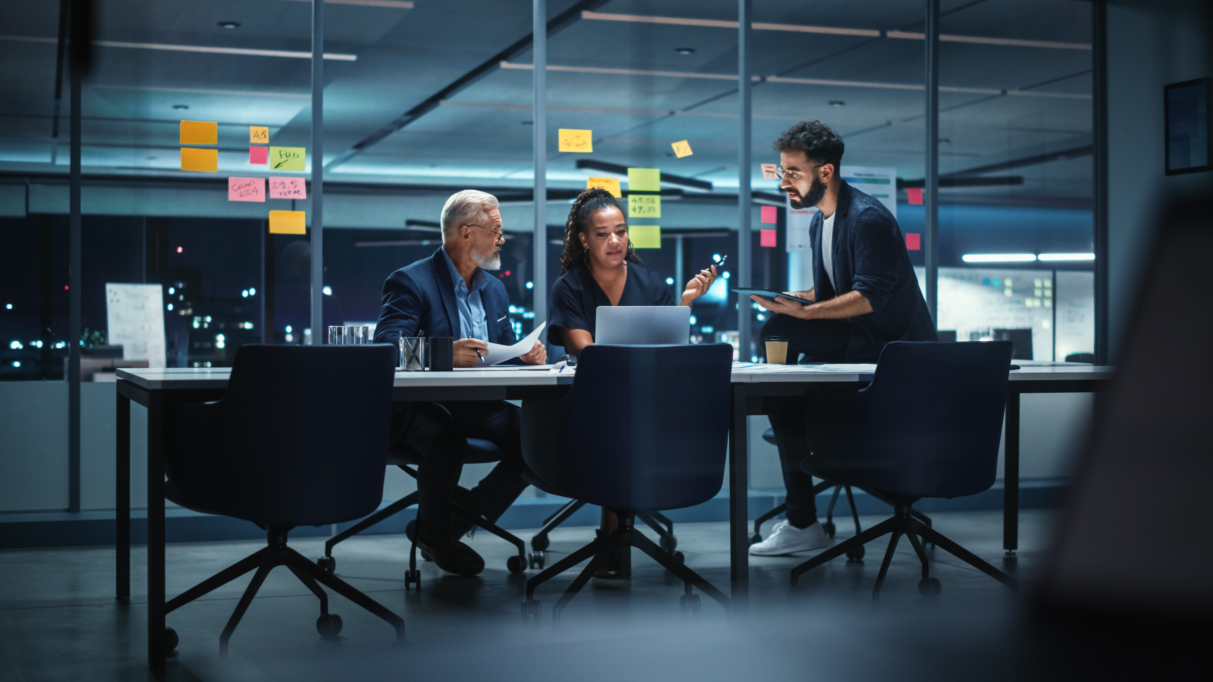 Three senior enterprise leaders review strategy documents and data late at night in a glass-walled conference room, representing executive decision-making in a fast-moving technology market.