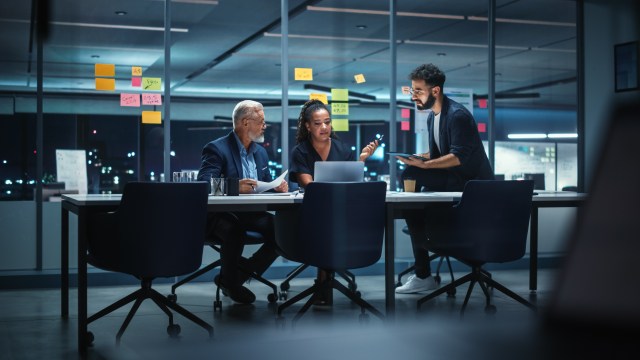 Three senior enterprise leaders review strategy documents and data late at night in a glass-walled conference room, representing executive decision-making in a fast-moving technology market.