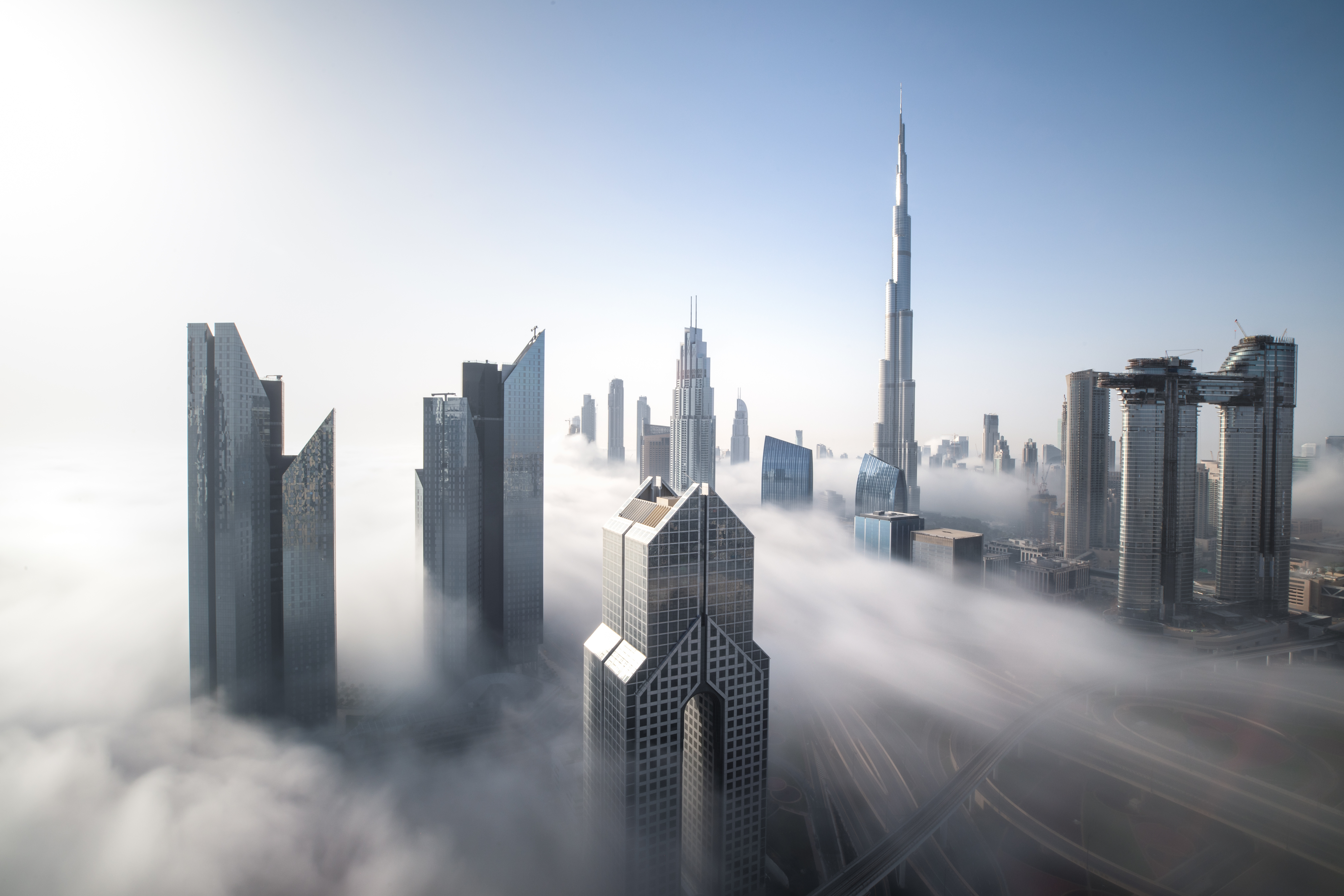 Dubai skyscrapers, including the Burj Khalifa, rising above a thick layer of fog under a clear blue sky