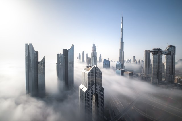 Dubai skyscrapers, including the Burj Khalifa, rising above a thick layer of fog under a clear blue sky