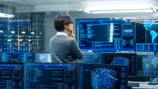 IT security operator in a control room monitoring multiple screens displaying network data, system analytics, and world map visualizations