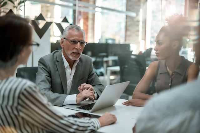 A senior IT leader discusses a decision with colleagues around a laptop in a modern office meeting
